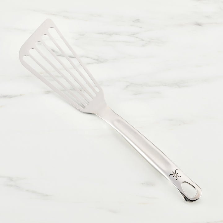 A Hestan Provisions Fish Turner, 13-inch stainless steel slotted spatula, essential for chefs, rests on a white marble surface. It features a long handle with a decorative cut-out near the end.