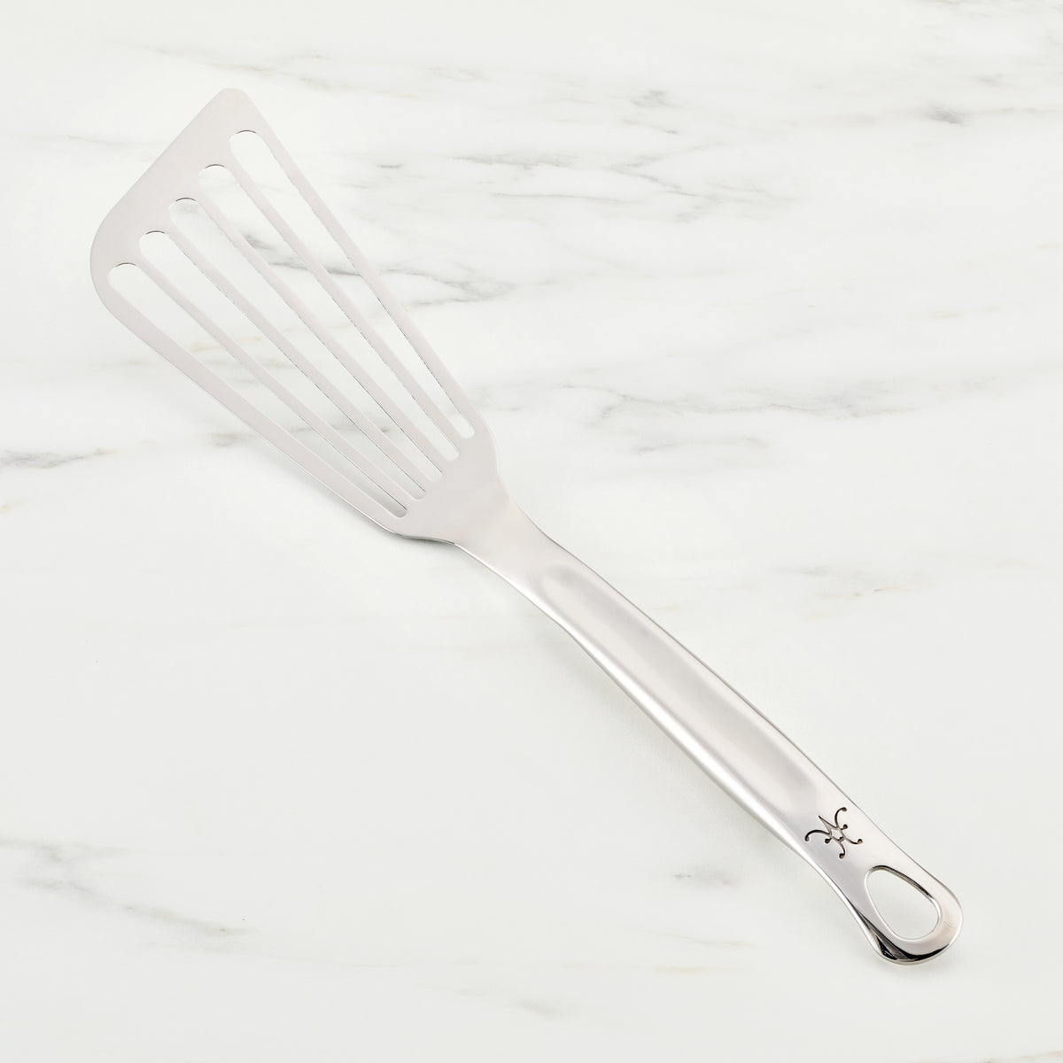 A Hestan Provisions Fish Turner, 13-inch stainless steel slotted spatula, essential for chefs, rests on a white marble surface. It features a long handle with a decorative cut-out near the end.