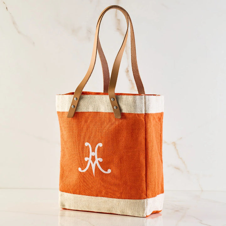 The Hestan Market Tote, Citra Orange by Accessories, is a masterful blend of orange and beige with a resilient design. It stands upright against a marble background, featuring brown leather straps and a white decorative symbol, embodying durable luxury.