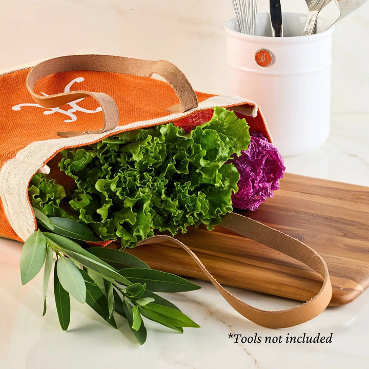 An open Hestan Market Tote in Citra Orange displays its durable design on a marble surface filled with lettuce and leafy branches. Nearby are a wooden cutting board and a white container with kitchen utensils. Text notes, *Tools not included. Brand: Accessories.