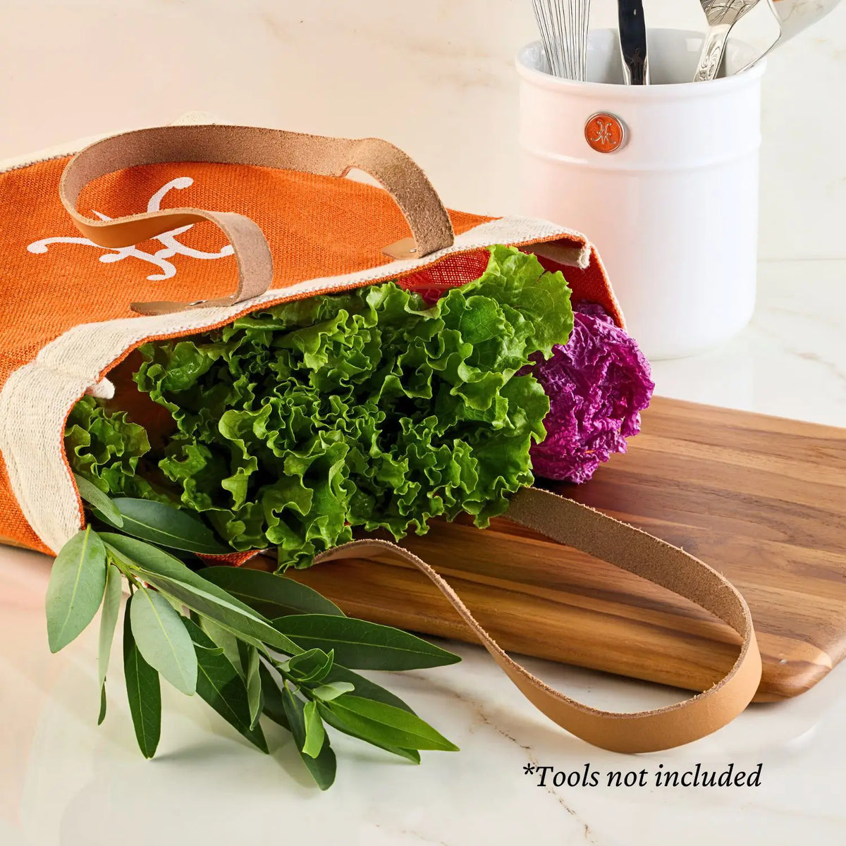 An open Hestan Market Tote in Citra Orange displays its durable design on a marble surface filled with lettuce and leafy branches. Nearby are a wooden cutting board and a white container with kitchen utensils. Text notes, *Tools not included. Brand: Accessories.