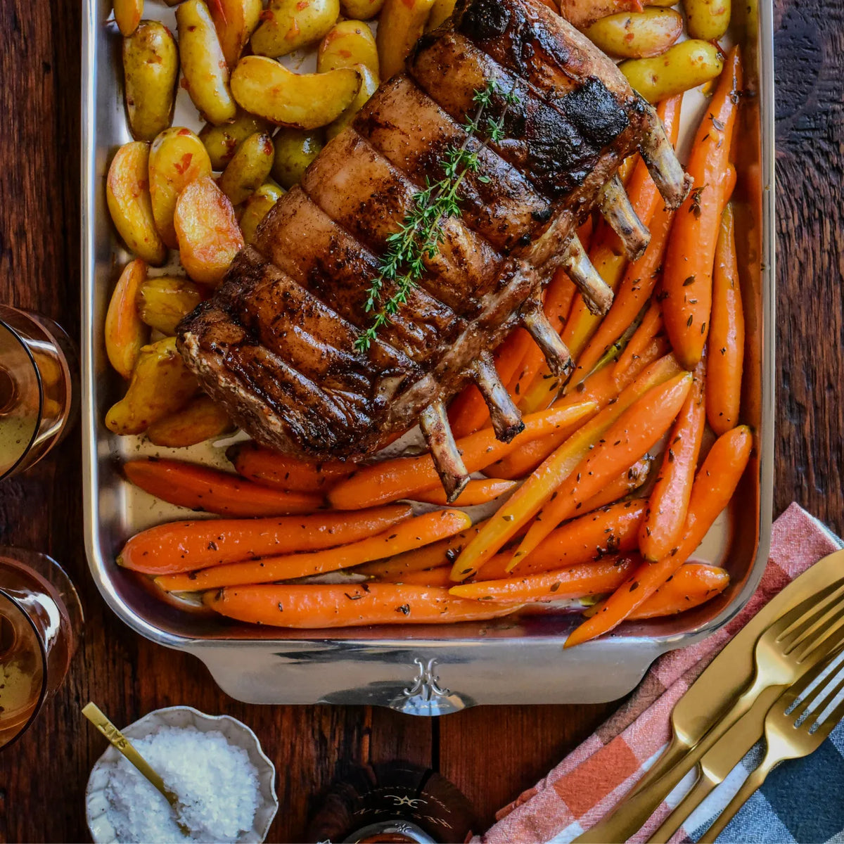 A roasted rack of pork ribs with herbs rests on the Hestan OvenBond Tri-ply Medium Sheet Pan with Rack, alongside roasted carrots and fingerling potatoes. Two glasses, a dish of salt, and cutlery sit on a wooden table nearby.