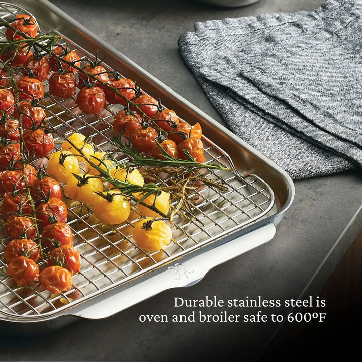 Roasted red and yellow tomatoes rest on a Hestan OvenBond Tri-Ply Medium Sheet Pan & Rack, 12" x 15", beside folded gray towels. Text: “Durable stainless steel is oven and broiler safe to 600°F.”.