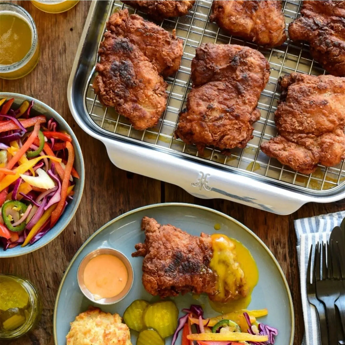 Fried chicken on a Hestan OvenBond OvenBond Tri-Ply Medium Sheet Pan & Rack, 12" x 15", next to a plate with fried chicken, pickles, coleslaw, a biscuit, sauces, and vegetable salad on a wooden table.