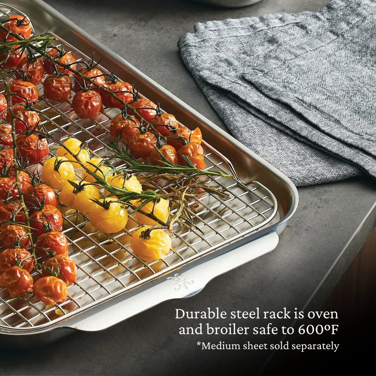 A Hestan OvenBond OvenBond Stainless Steel Medium Pan Rack holds roasted yellow and red tomatoes on the vine atop a baking sheet. Gray oven mitts are nearby. Text: Durable rack is oven and broiler safe to 600°F. *Medium sheet sold separately.