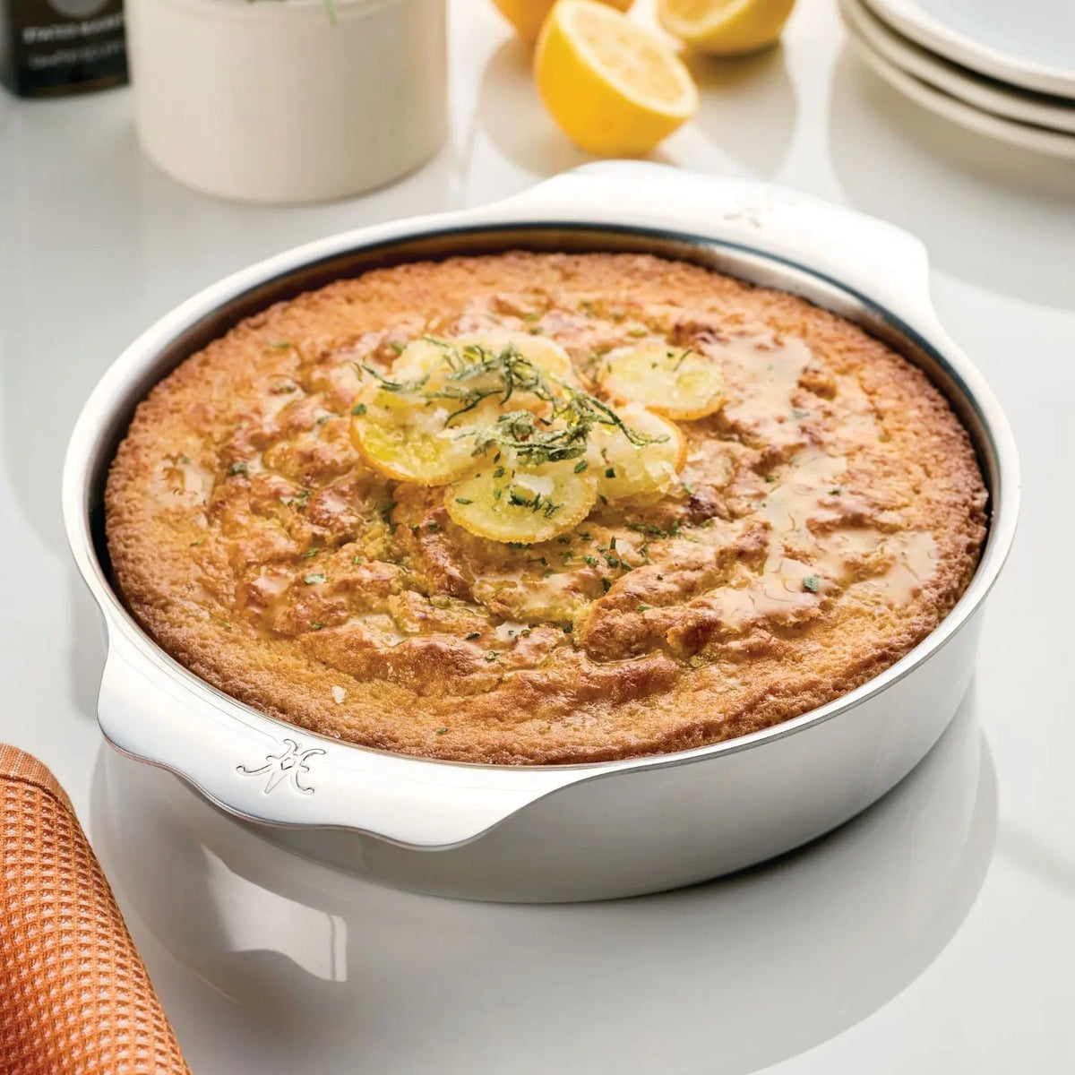 A lemon cake with lemon slices and herbs rests in the Hestan OvenBond Round Baker, 9-inch, on a white surface. Plates, lemon halves, and a plant are visible in the background.