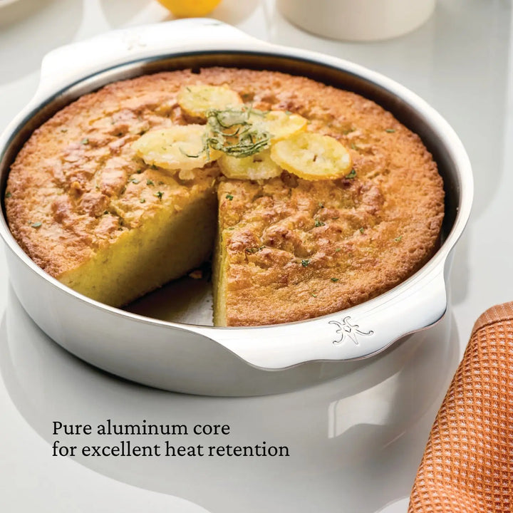 A round cake with a slice missing sits in a silver Hestan OvenBond OvenBond Round Baker, 9-inch. Lemon slices and herbs top the cake. Black text reads: Pure aluminum core for excellent heat retention in this tri-ply cake pan.