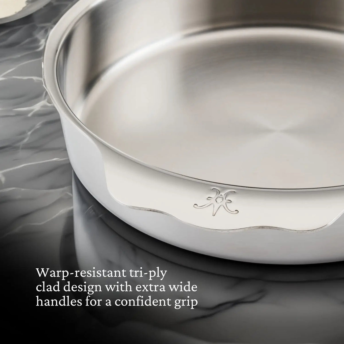 A close-up of the Hestan OvenBond Round Baker, 9-inch, shows its tri-ply clad stainless steel design and extra wide handles on a dark marble countertop. Text highlights warp resistance and sturdy grip—perfect for commercial bakeware needs.