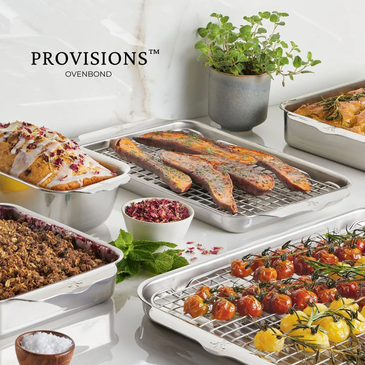 A Hestan OvenBond 9-inch Round Baker sits on the kitchen countertop, filled with roasted tomatoes, sausages, a loaf with icing, and baked granola beside a potted plant and a bowl of coarse salt.
