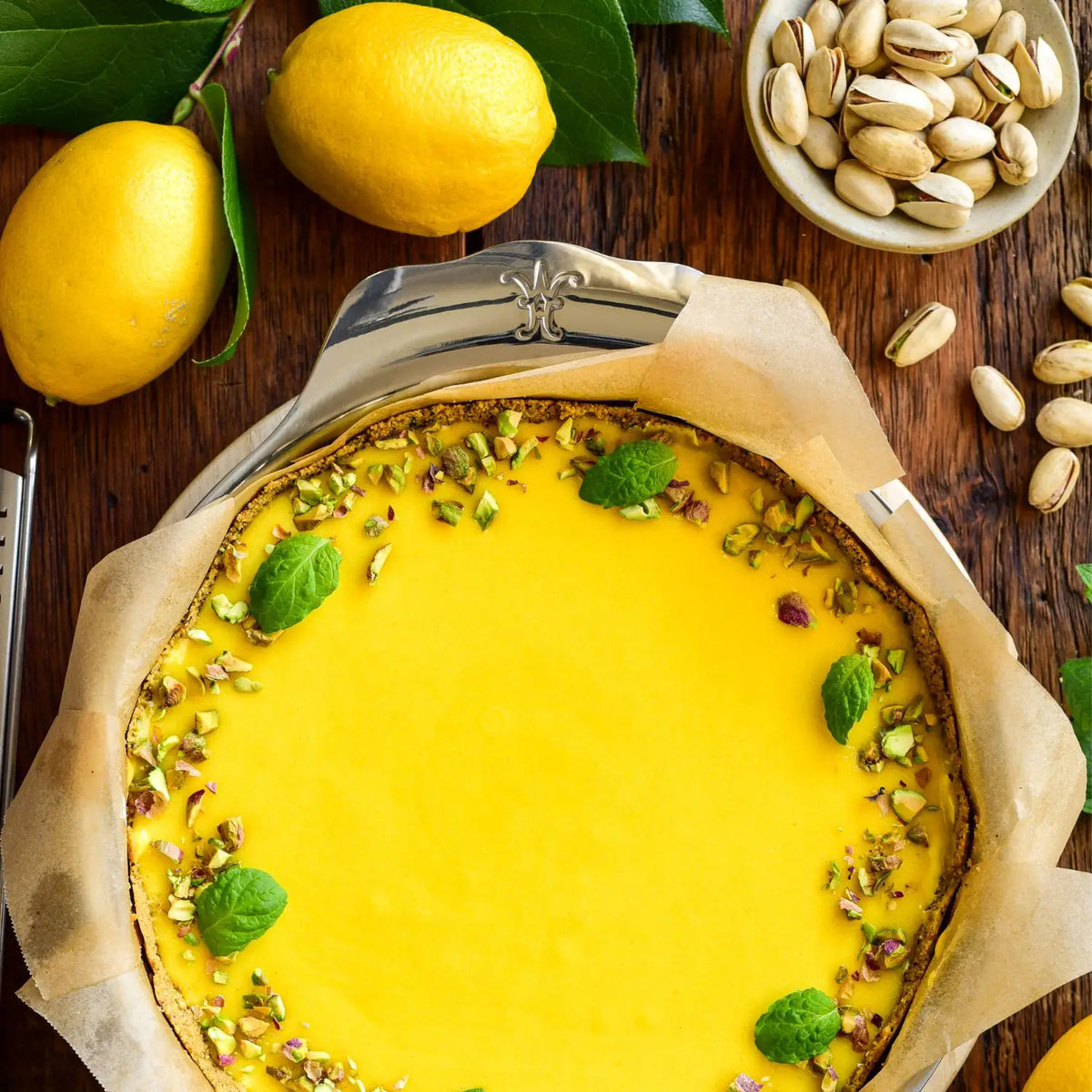 A freshly baked lemon tart sits on parchment in the Hestan OvenBond Round Baker, 9-inch, topped with pistachios and mint. Whole lemons, green leaves, and pistachios are displayed nearby on a wooden surface.
