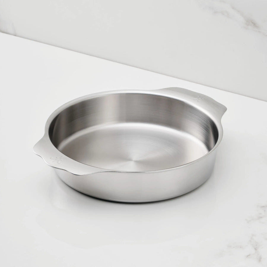 The Hestan OvenBond Round Baker, 9-inch, with its tri-ply design and two flat handles, sits on a marble-patterned white countertop, highlighting the durability and elegance of quality commercial bakeware.