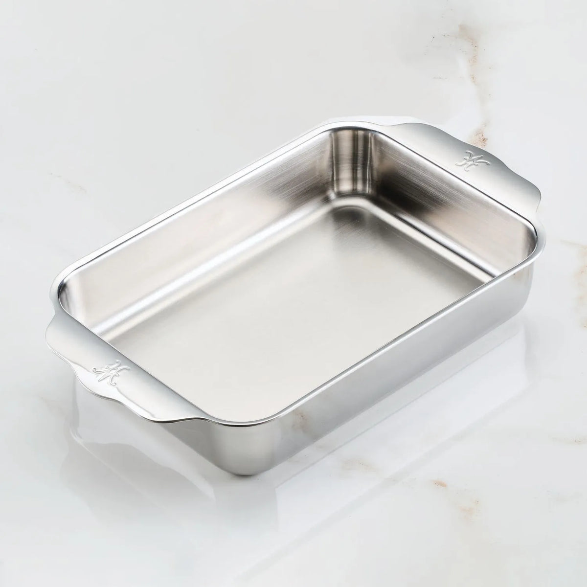 The Hestan OvenBond Tri-ply Small Rectangular Baker features wide handles, a brushed stainless steel interior, a shiny exterior, and is oven safe up to 600°F. It’s shown resting on a white marble surface.