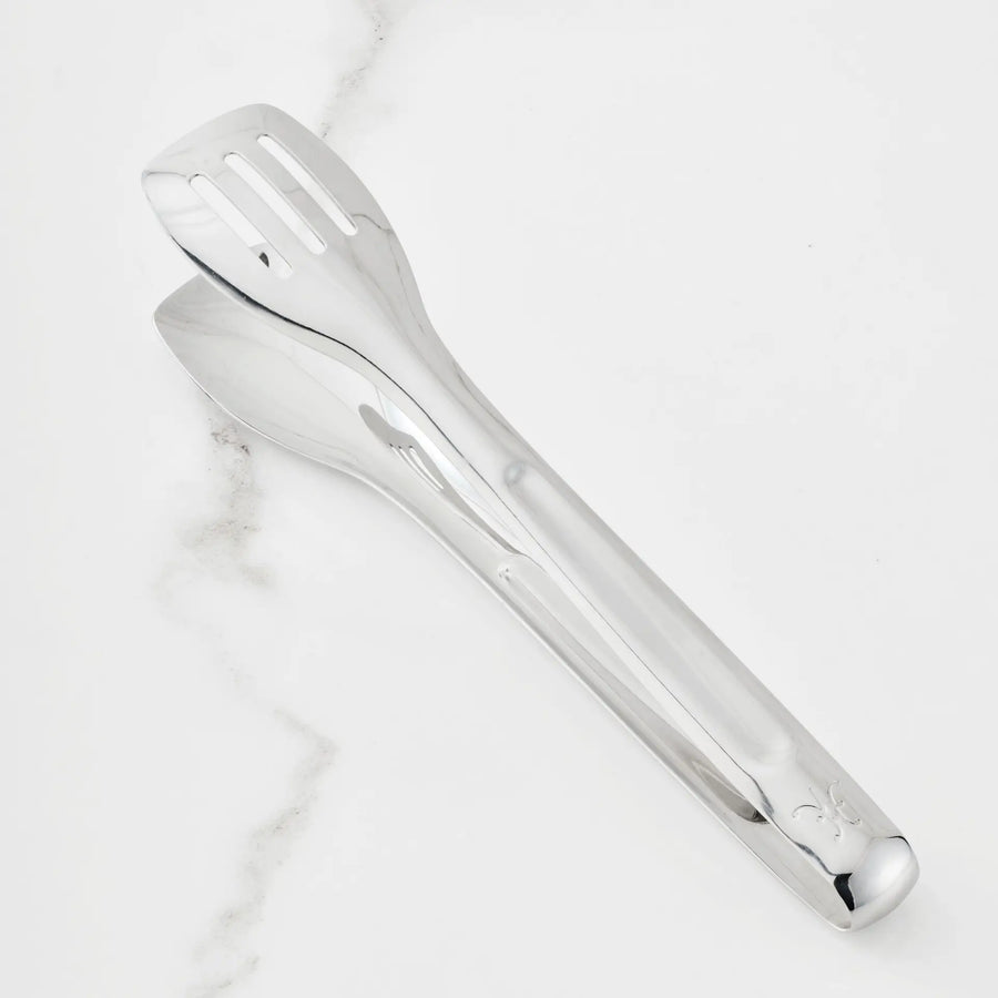 A pair of 10-inch Stainless Steel Serving Tongs from the Hestan Provisions® collection rests on a white marble surface. One side features a slotted, flat end while the other has a spoon-shaped end, and the handle is finished with a small embossed design near the tip—an elegant addition to any chef’s kitchen tools.