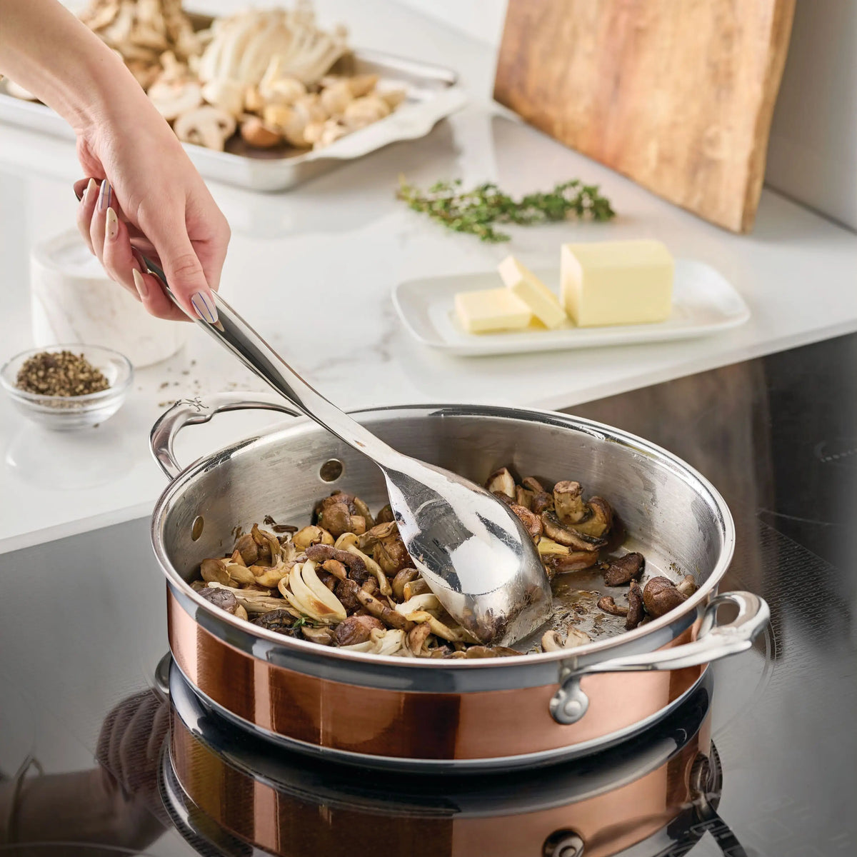 A hand uses the Hestan Provisions Sauté Spoon with Straight Edge, 13-inch, to stir sliced mushrooms and onions in a copper pan on an electric stove. Butter, herbs, uncooked mushrooms, and a small bowl of pepper are nearby.