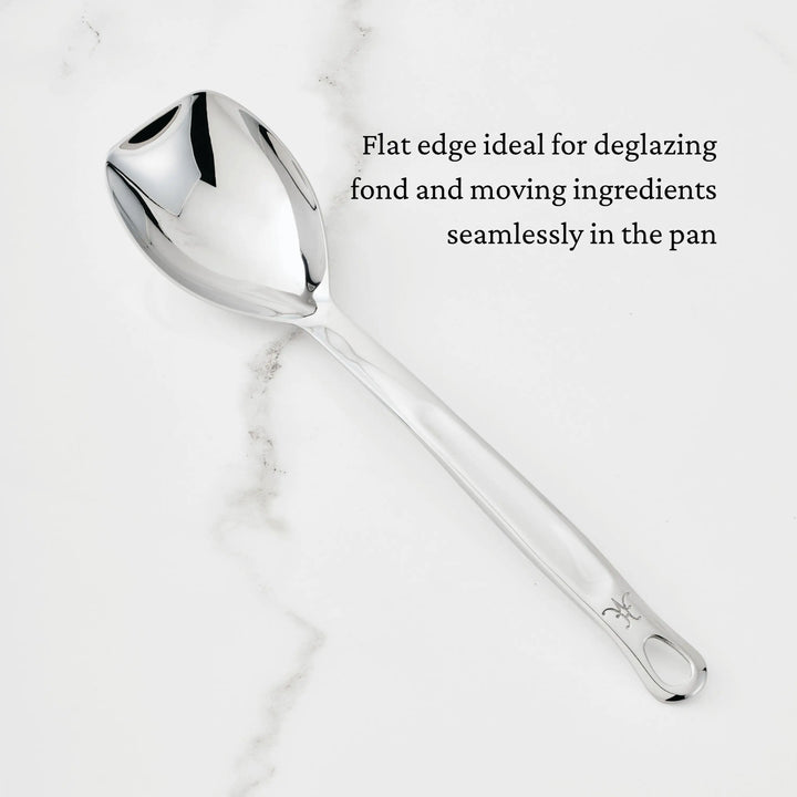 A Hestan Provisions Sauté Spoon with Straight Edge, 13-inch, rests on white marble. Black text reads, “Flat edge ideal for deglazing fond and moving ingredients seamlessly in the pan.”.