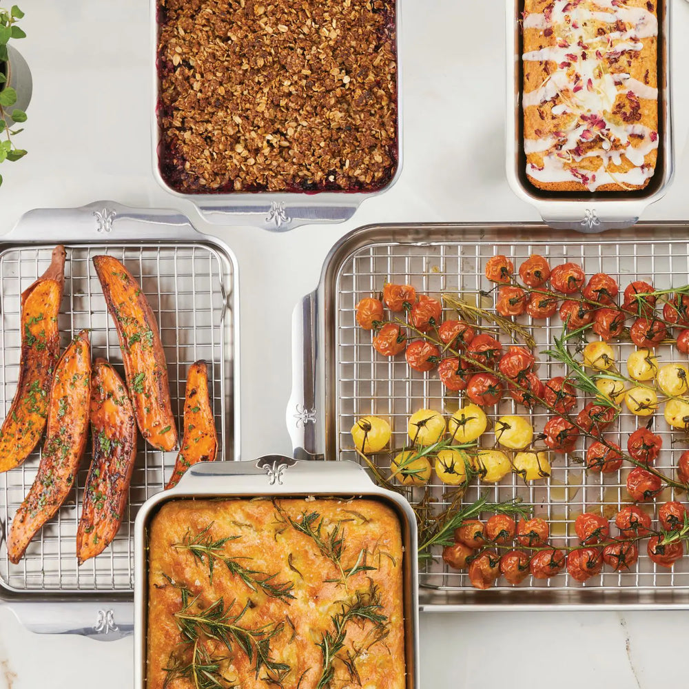 Five trays of food, each artfully arranged in Hestan OvenBond Tri-ply Gourmet Essentials Stainless Steel bakeware—featuring sweet potato wedges, veggie skewers, rosemary focaccia, an iced dessert, and fruit crumble.