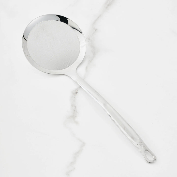 A Hestan Provisions Fine Mesh Skimmer, 16-inch, with a long handle and stainless steel head rests on a white marble surface.