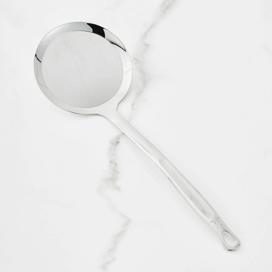 A Hestan Provisions Fine Mesh Skimmer, 16-inch, with a long handle and stainless steel head rests on a white marble surface.