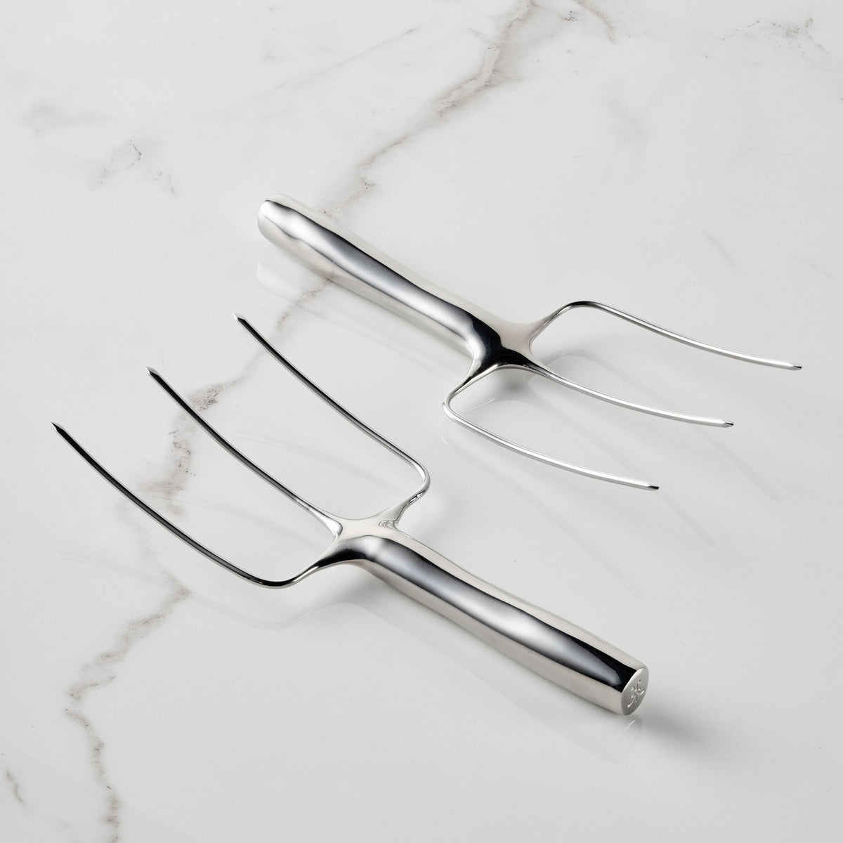 Two Hestan Provisions® Stainless Steel Carving Forks, Set of 2—each featuring three prongs and crafted from durable 18/10 stainless steel—are displayed on a white marble surface, with one fork facing up and the other facing down.