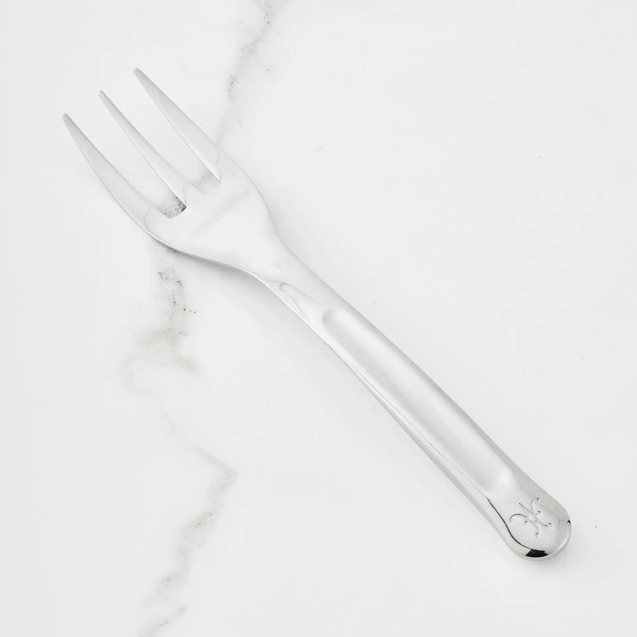 A 10.5-inch Stainless Steel Serving Fork from Hestan Provisions® rests on a white marble surface. Featuring three wide tines and a handle with a small engraved design near the end, this serving fork is an essential addition to any chef’s toolkit.