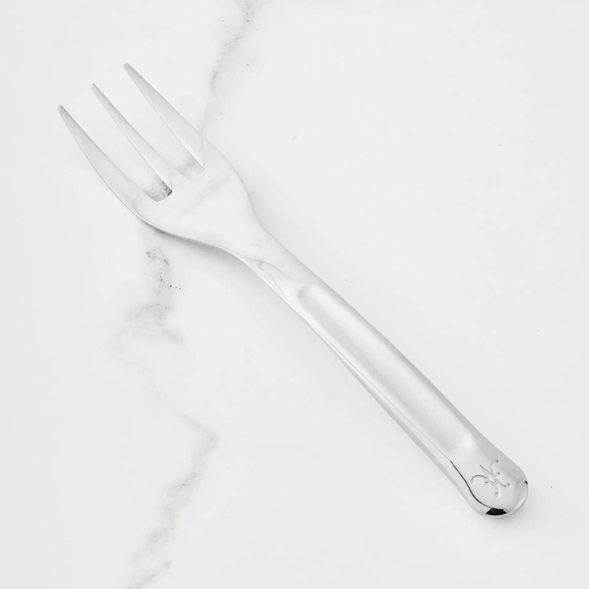 A 10.5-inch Stainless Steel Serving Fork from Hestan Provisions® rests on a white marble surface. Featuring three wide tines and a handle with a small engraved design near the end, this serving fork is an essential addition to any chef’s toolkit.