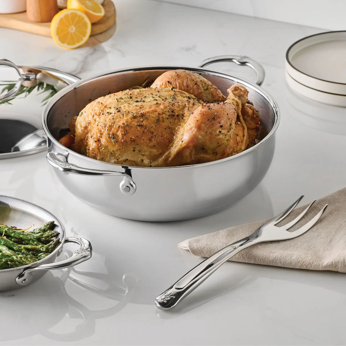 A whole roasted chicken rests in a round stainless steel pot with handles. Nearby is the Hestan Provisions® 10.5-inch Stainless Steel Serving Fork placed on a napkin, accompanied by a dish of green beans, stacked plates, and lemon slices on a cutting board—everything you need among your chef’s tools for an inviting meal.