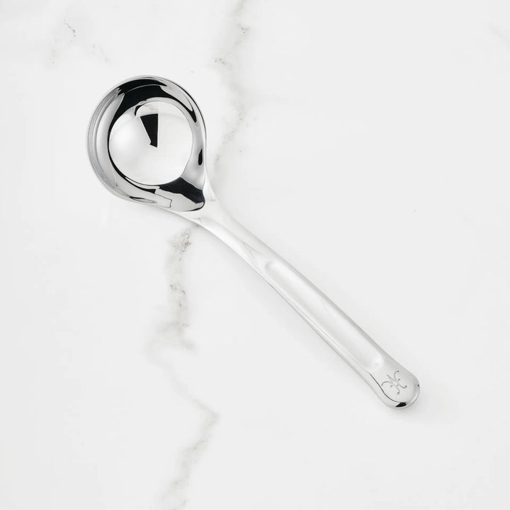 The 2.4-oz Stainless Steel Serving Ladle from Hestan Provisions®—featuring a round bowl, long handle with a small decorative mark at the end—is placed on a white marble surface.