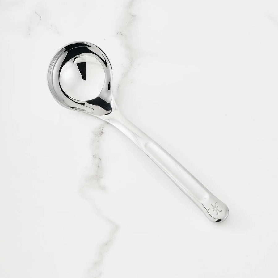 The 2.4-oz Stainless Steel Serving Ladle from Hestan Provisions®—featuring a round bowl, long handle with a small decorative mark at the end—is placed on a white marble surface.