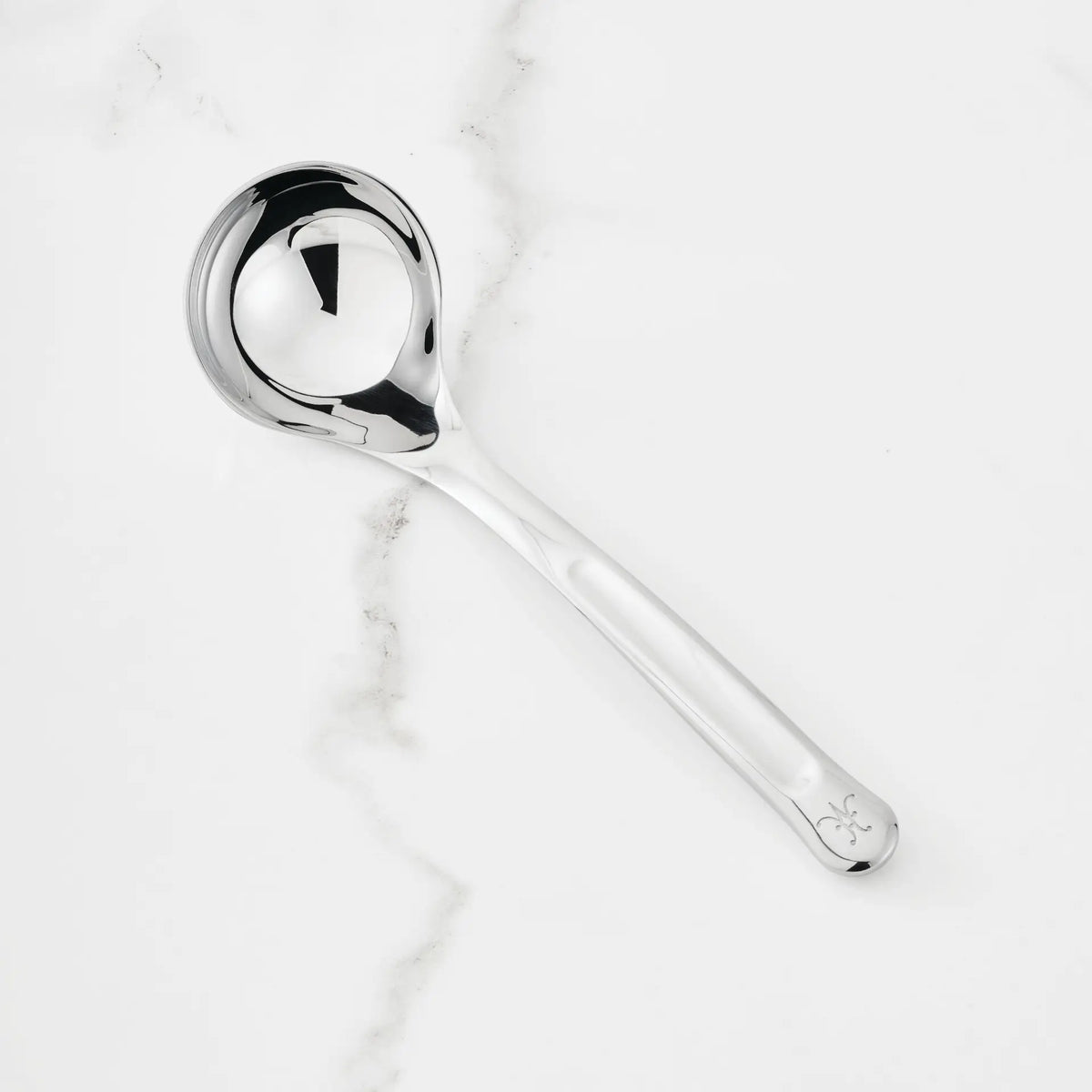 The 2.4-oz Stainless Steel Serving Ladle from Hestan Provisions®—featuring a round bowl, long handle with a small decorative mark at the end—is placed on a white marble surface.