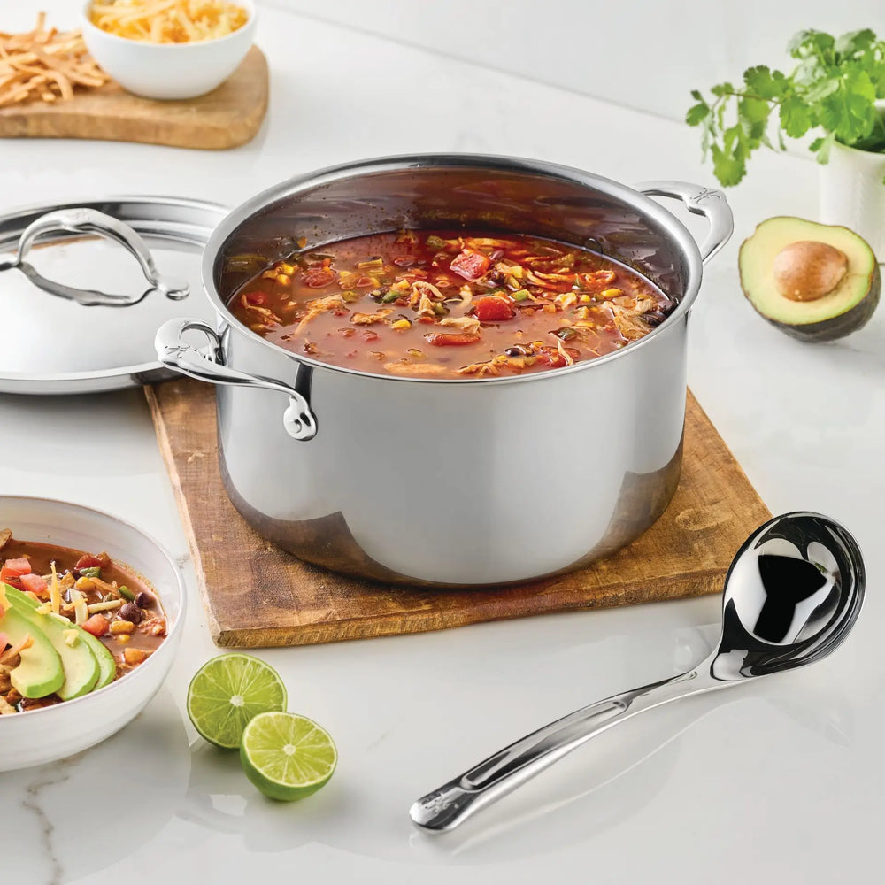 A stainless steel pot filled with soup rests on a wooden board, accompanied by the Hestan Provisions® 2.4-oz Stainless Steel Serving Ladle. Sliced limes, a bowl of soup, avocado, cilantro, and bread are artfully arranged nearby on a white countertop, highlighting the elegance of stainless steel kitchen tools from Hestan Provisions® in a modern kitchen setting.