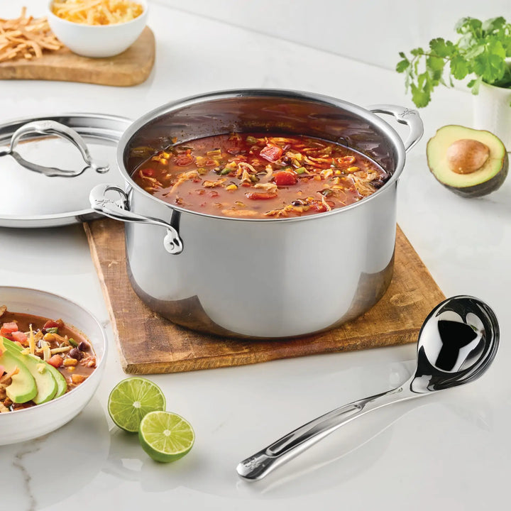 A stainless steel pot filled with soup rests on a wooden board, accompanied by the Hestan Provisions® 2.4-oz Stainless Steel Serving Ladle. Sliced limes, a bowl of soup, avocado, cilantro, and bread are artfully arranged nearby on a white countertop, highlighting the elegance of stainless steel kitchen tools from Hestan Provisions® in a modern kitchen setting.
