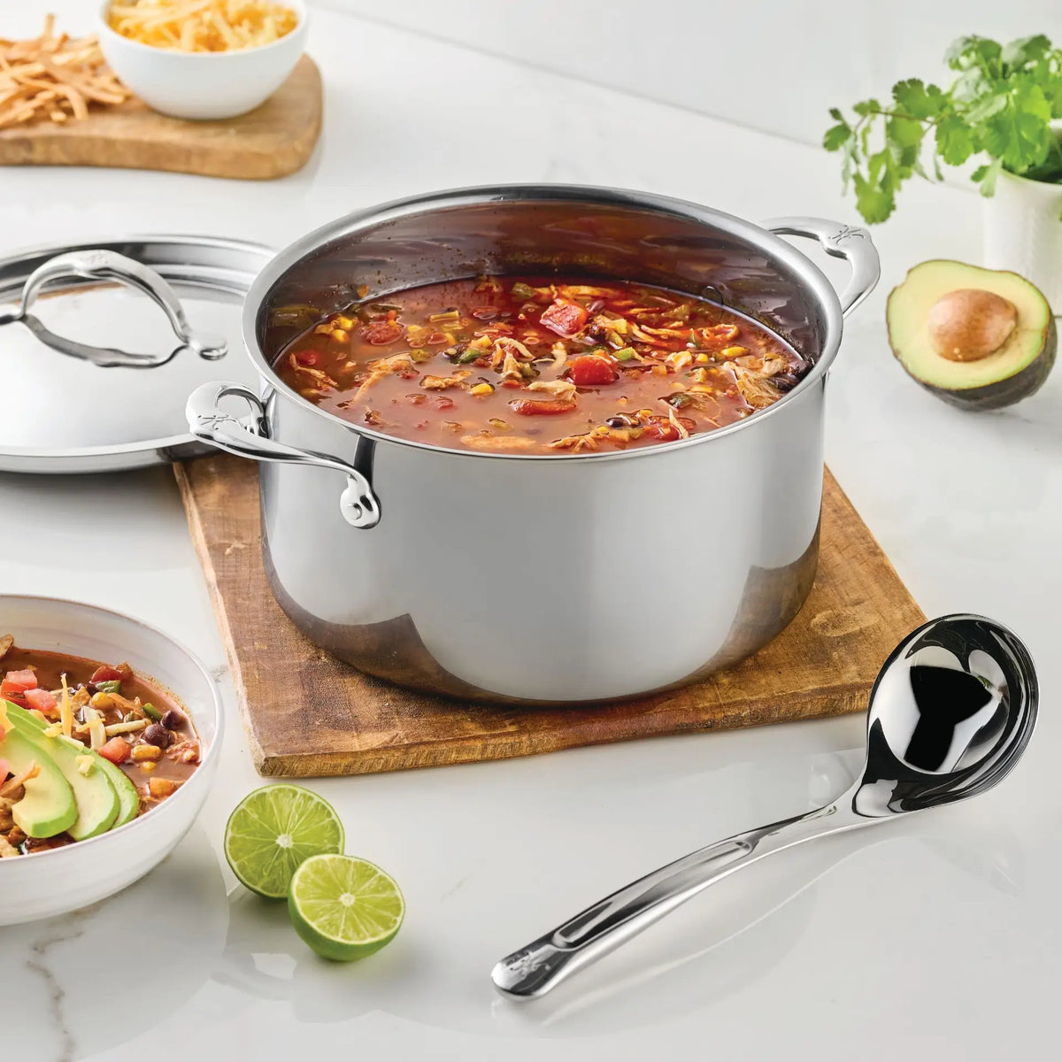 A stainless steel pot filled with soup rests on a wooden board, accompanied by the Hestan Provisions® 2.4-oz Stainless Steel Serving Ladle. Sliced limes, a bowl of soup, avocado, cilantro, and bread are artfully arranged nearby on a white countertop, highlighting the elegance of stainless steel kitchen tools from Hestan Provisions® in a modern kitchen setting.