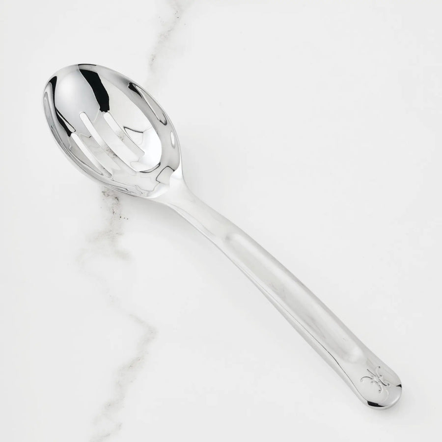 The 10.5-inch Stainless Steel Slotted Serving Spoon from Hestan Provisions® rests on a white marble countertop, its reflective surface gleaming.