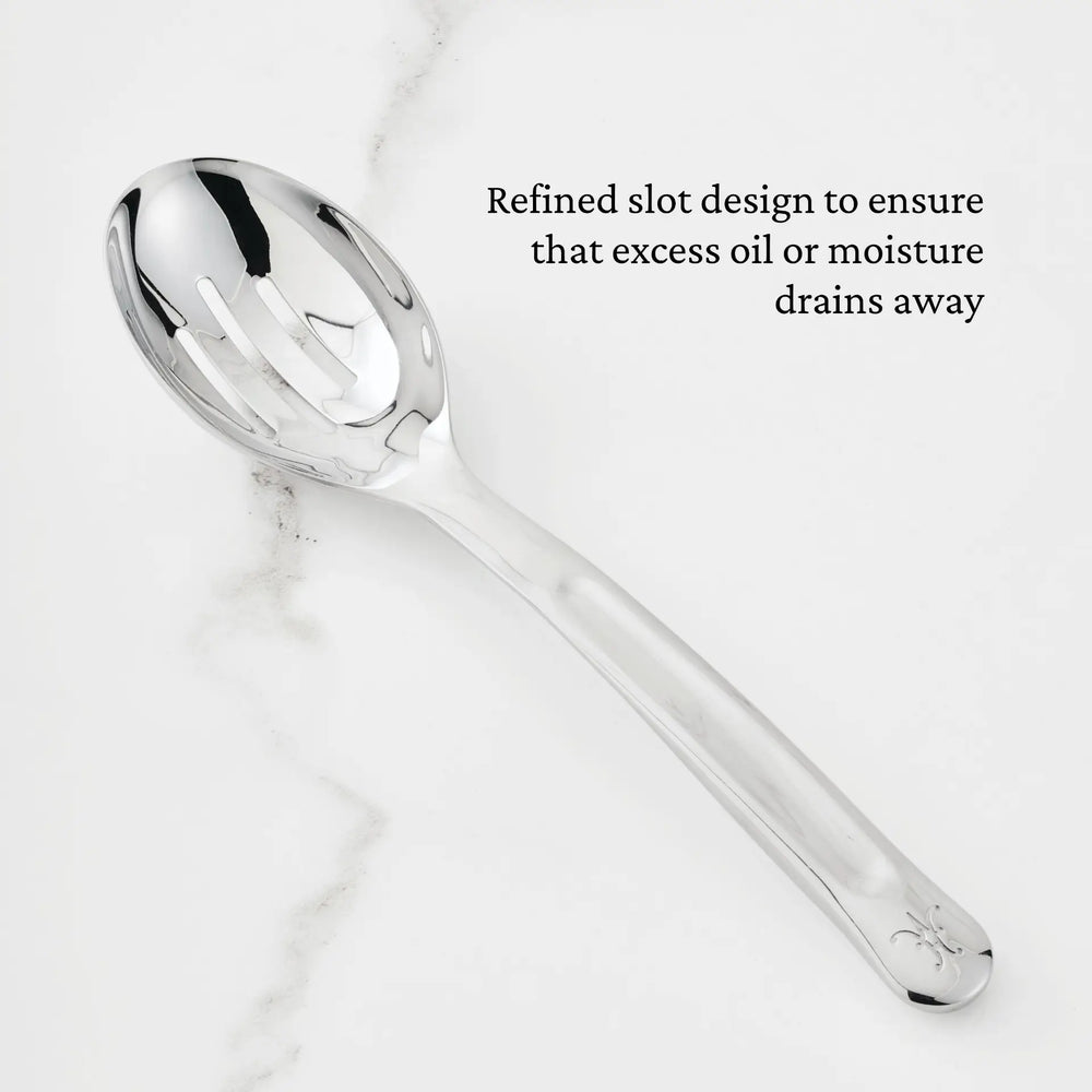 A 10.5-inch Stainless Steel Slotted Serving Spoon from Hestan Provisions® rests on a white marble surface. Black text beside the spoon reads, Refined slot design to ensure that excess oil or moisture drains away.