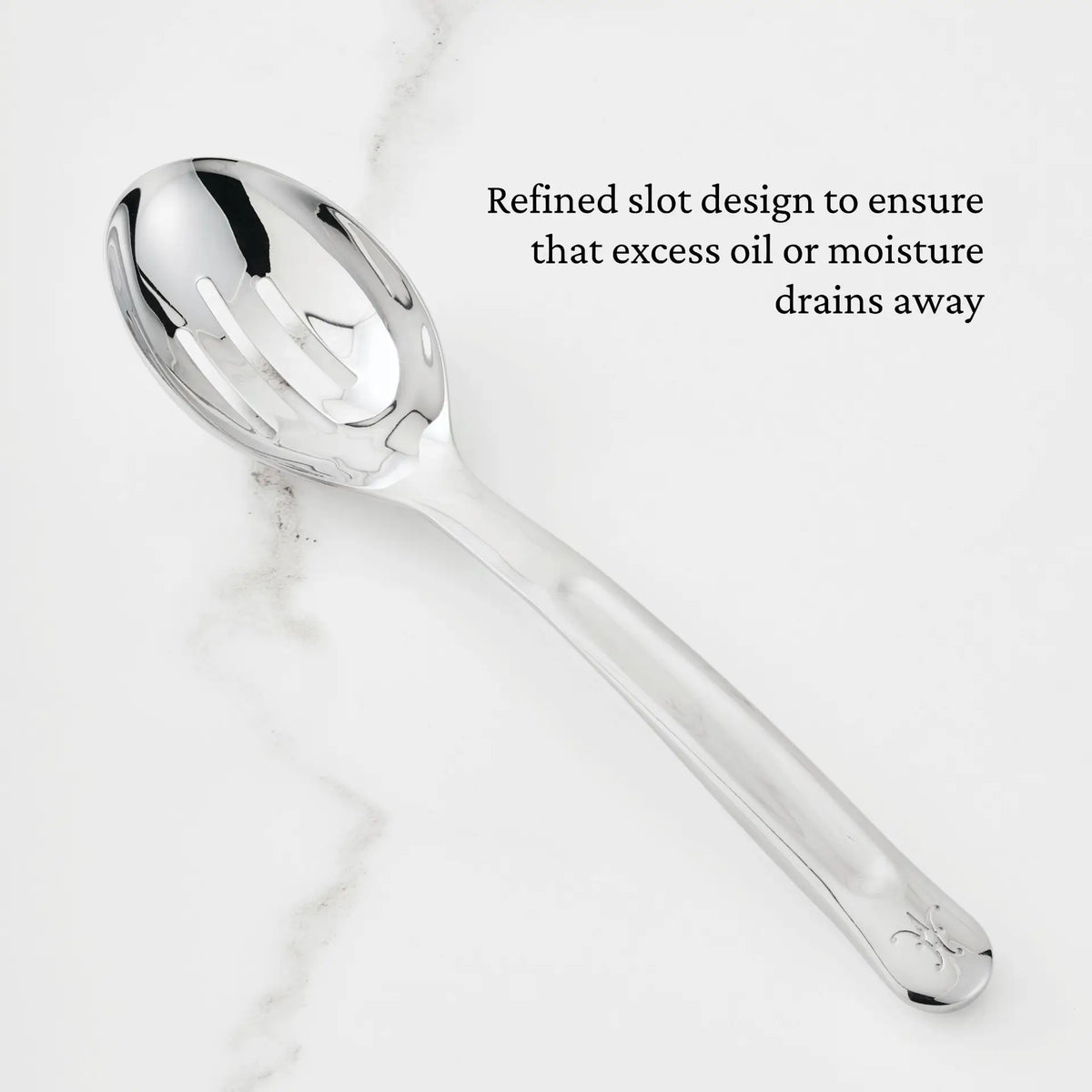 A 10.5-inch Stainless Steel Slotted Serving Spoon from Hestan Provisions® rests on a white marble surface. Black text beside the spoon reads, Refined slot design to ensure that excess oil or moisture drains away.