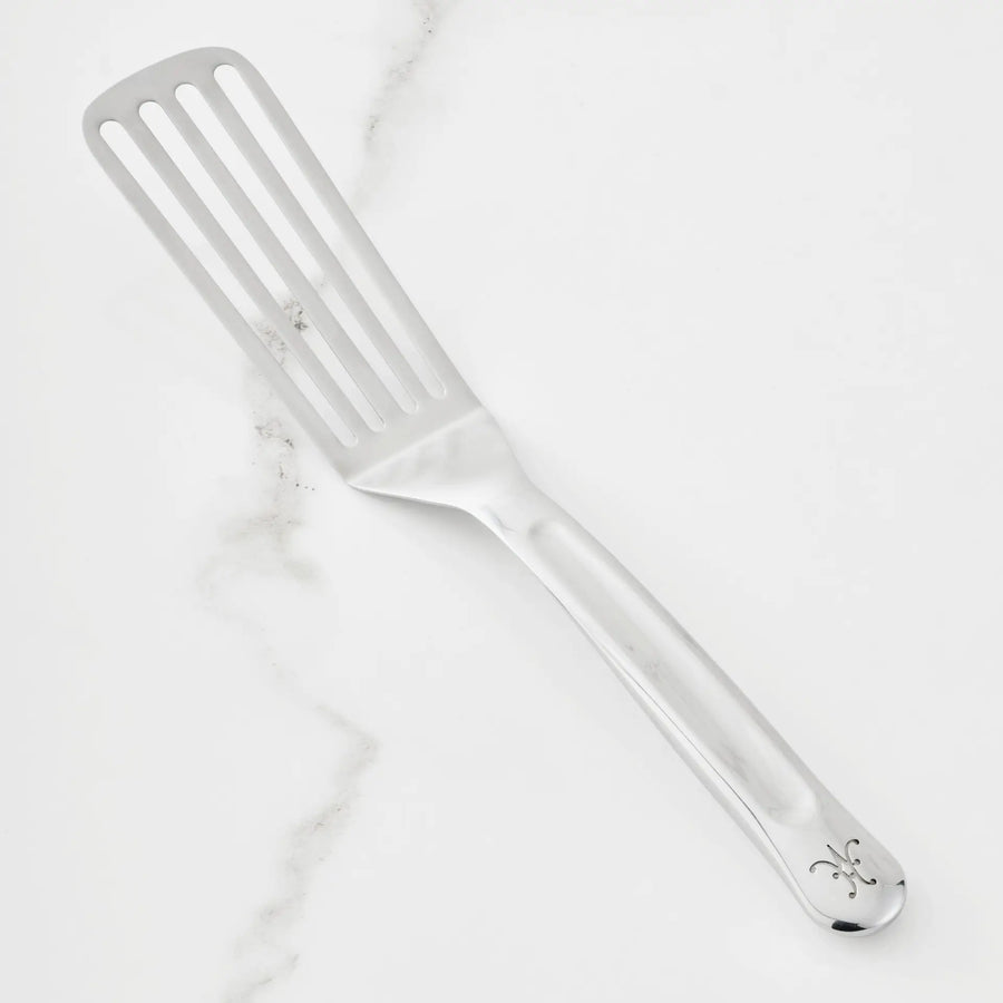 An 11.5-inch Stainless Steel Chef’s Fish Turner from Hestan Provisions®, featuring a long handle with decorative engraving near the end, is displayed on a white marble surface.