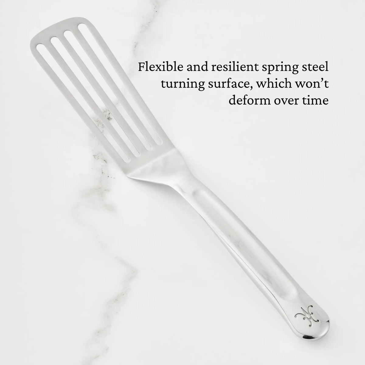 An 11.5-inch Stainless Steel Chef’s Fish Turner from Hestan Provisions® rests on a white marble surface. In black text above, it’s described as flexible and resilient, crafted from spring steel that resists deformation over time—ideal for those looking for professional-quality kitchen utensils.