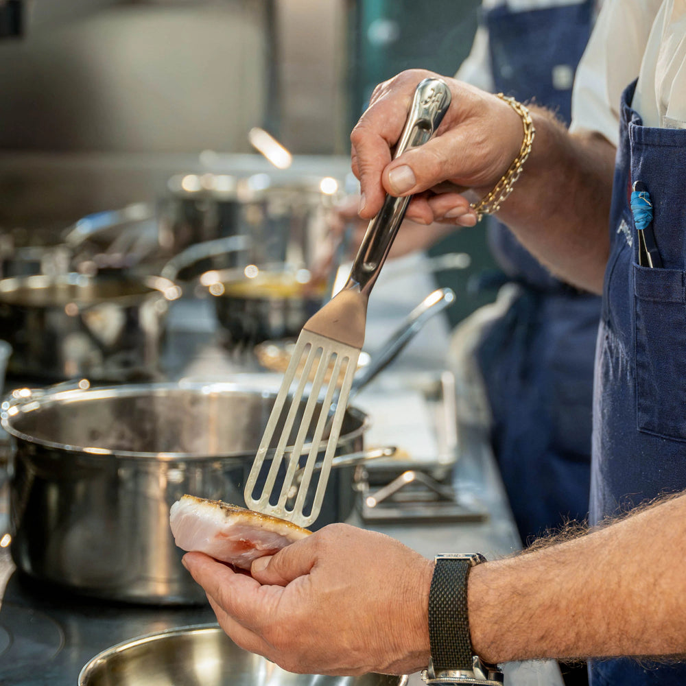 A person wearing a blue apron holds the Hestan Provisions® 11.5-inch Stainless Steel Chef’s Fish Turner and a piece of fish in a kitchen, preparing to cook it in a pan on the stove. Professional kitchen utensils and stainless steel pots, including Hestan Chef’s Tools, are visible in the background.