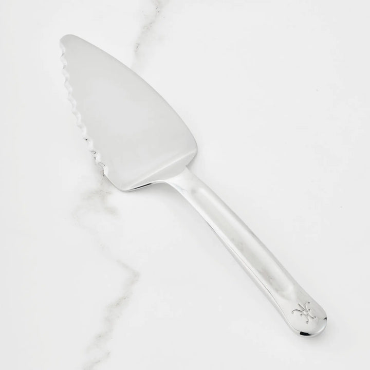 A 10.5-inch Stainless Steel Pie & Cake Server from the Hestan Provisions® collection rests on a white marble surface. Featuring a serrated edge, this serving tool is adorned with a small decorative symbol near the end of its handle.