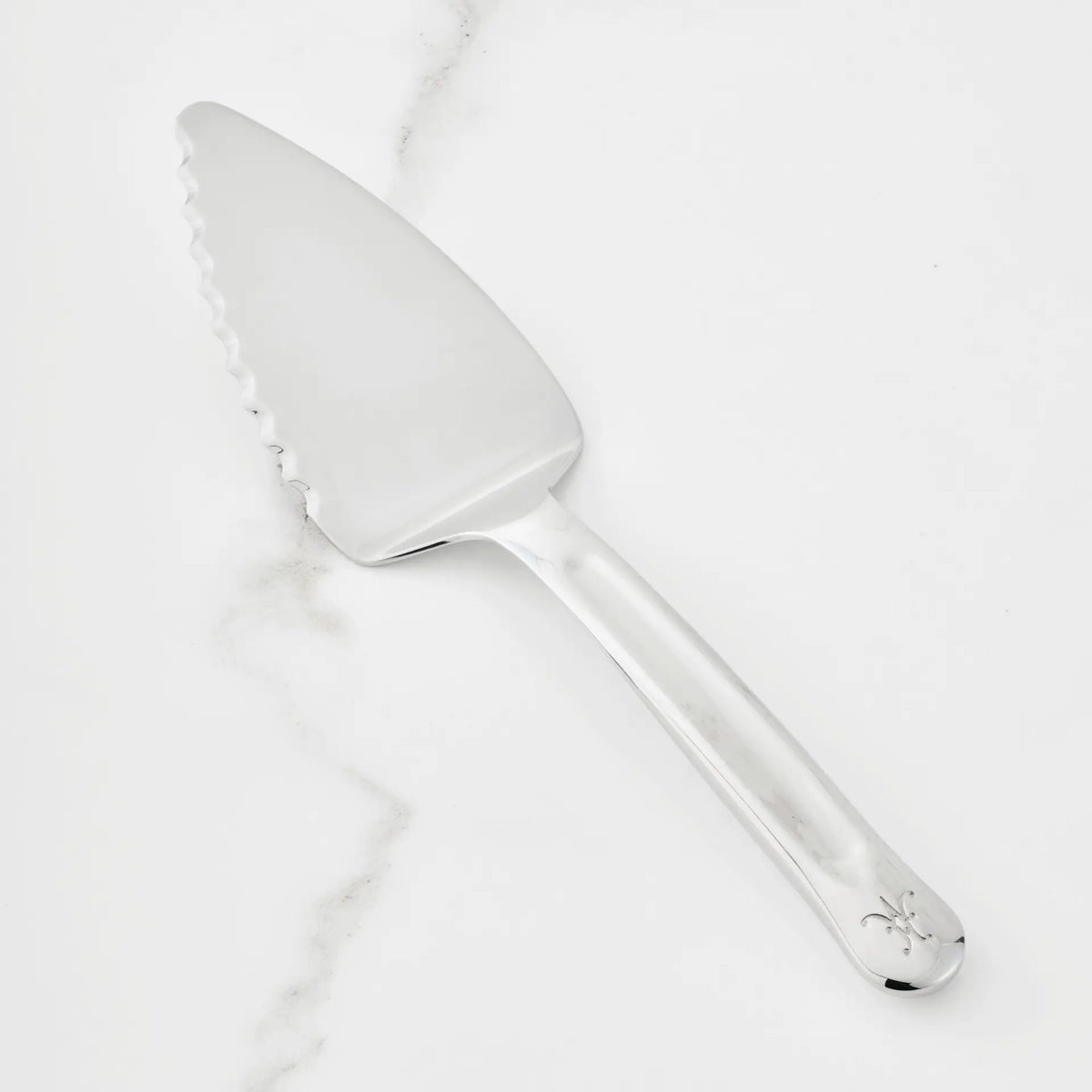 A 10.5-inch Stainless Steel Pie & Cake Server from the Hestan Provisions® collection rests on a white marble surface. Featuring a serrated edge, this serving tool is adorned with a small decorative symbol near the end of its handle.