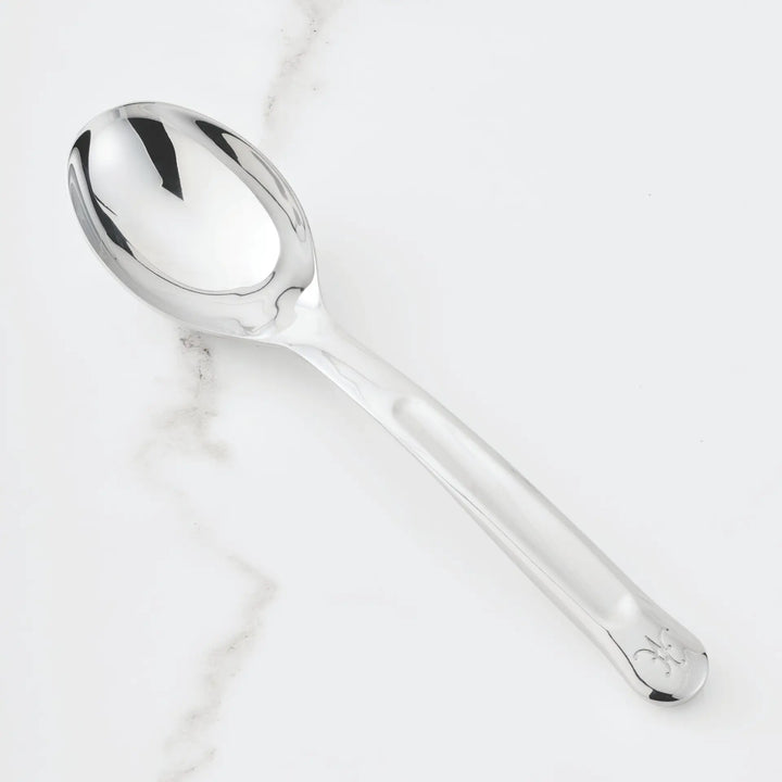 A gleaming 10.5-inch Stainless Steel Solid Serving Spoon from Hestan Provisions® rests on a white marble surface with delicate gray veining. Part of the Hestan Provisions® Serving Tools collection, this spoon features a sleek, reflective design with a subtle engraving near the end of its handle—an ideal choice for anyone who appreciates elegant chef’s tools in their kitchen.
