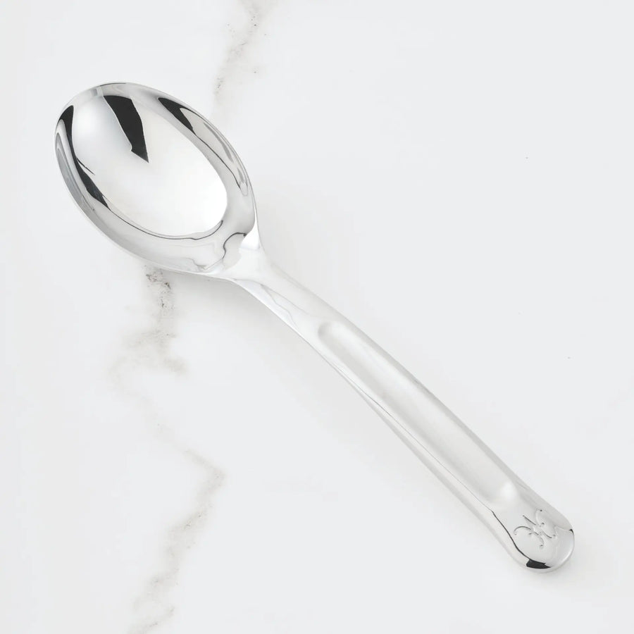 A gleaming 10.5-inch Stainless Steel Solid Serving Spoon from Hestan Provisions® rests on a white marble surface with delicate gray veining. Part of the Hestan Provisions® Serving Tools collection, this spoon features a sleek, reflective design with a subtle engraving near the end of its handle—an ideal choice for anyone who appreciates elegant chef’s tools in their kitchen.