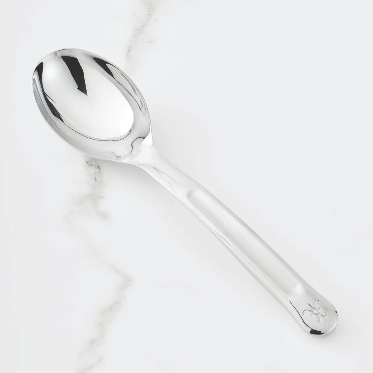 A gleaming 10.5-inch Stainless Steel Solid Serving Spoon from Hestan Provisions® rests on a white marble surface with delicate gray veining. Part of the Hestan Provisions® Serving Tools collection, this spoon features a sleek, reflective design with a subtle engraving near the end of its handle—an ideal choice for anyone who appreciates elegant chef’s tools in their kitchen.