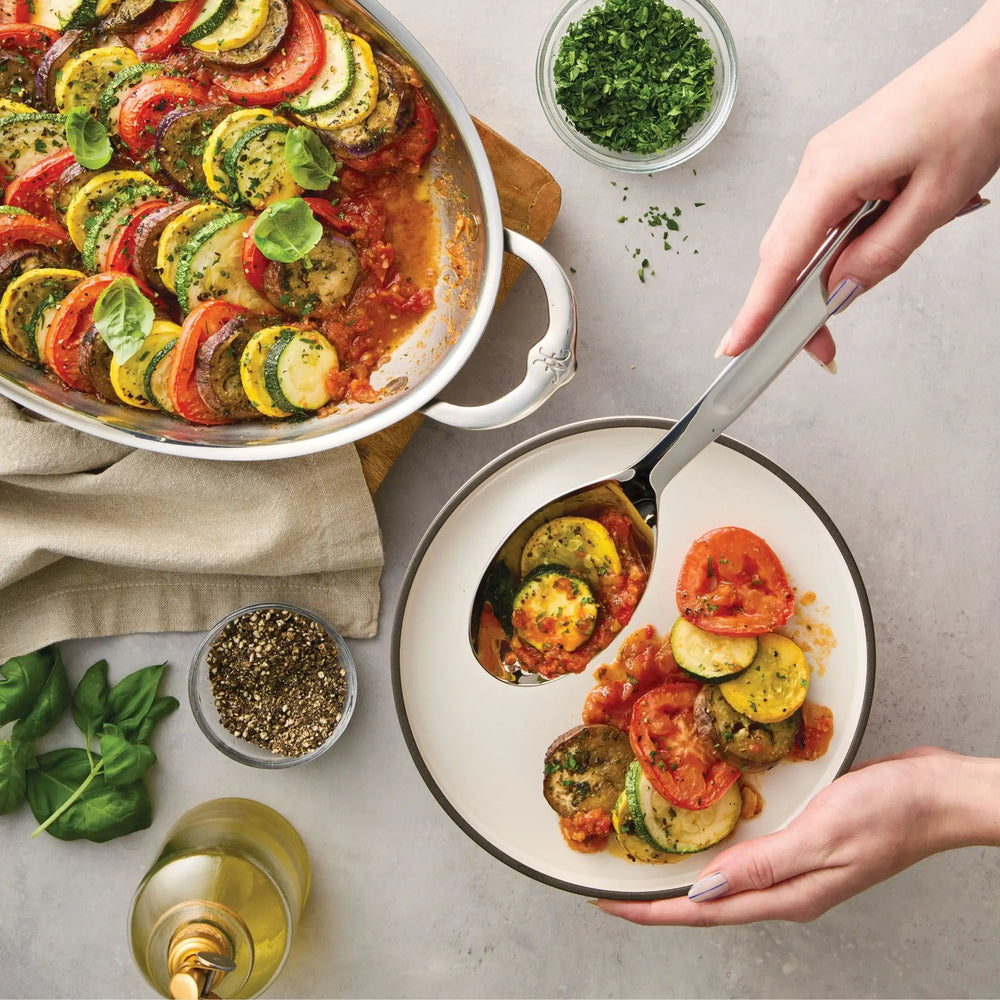 Using a Hestan Provisions® 10.5-inch Stainless Steel Solid Serving Spoon, a person serves sliced ratatouille from a white baking dish into a bowl. Fresh herbs, black pepper, basil leaves, and a glass of white wine are arranged on the gray tabletop nearby.