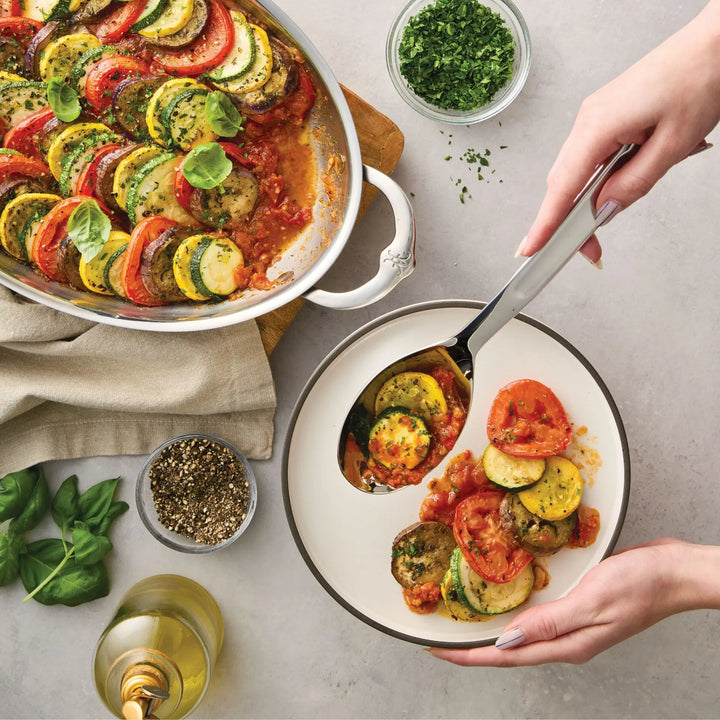 Using a Hestan Provisions® 10.5-inch Stainless Steel Solid Serving Spoon, a person serves sliced ratatouille from a white baking dish into a bowl. Fresh herbs, black pepper, basil leaves, and a glass of white wine are arranged on the gray tabletop nearby.