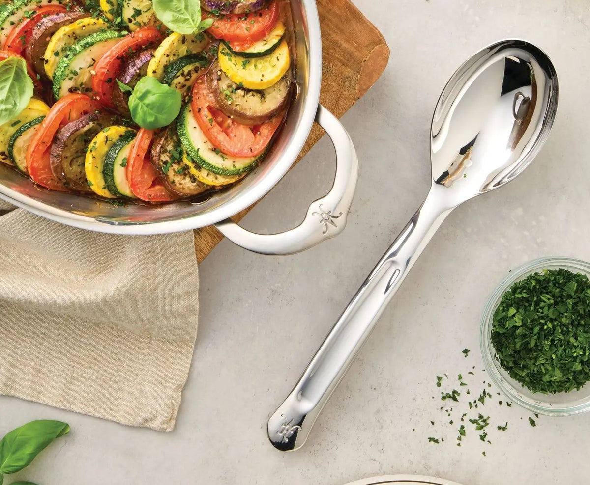 A casserole dish with sliced vegetables arranged in a spiral sits on a beige cloth next to a Hestan Provisions® 10.5-inch Stainless Steel Solid Serving Spoon and a small bowl of chopped herbs on a light countertop, showcasing the perfect blend of elegant presentation and premium chef’s tools.