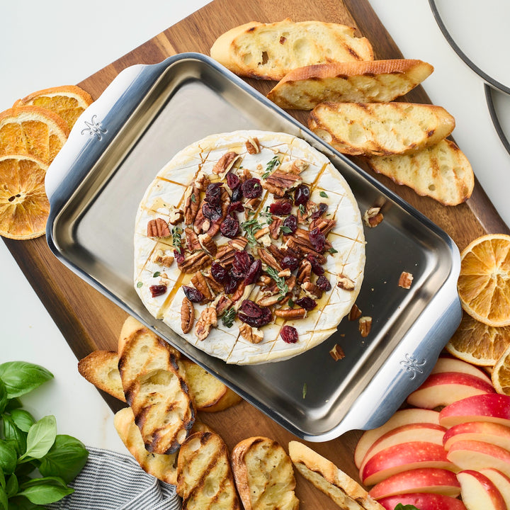 A round brie topped with pecans, cranberries, and herbs rests on the Hestan OvenBond Tri-ply Mini Eighth Sheet Pan with Rack, surrounded by grilled bread, apple wedges, dried orange slices, and fresh basil on a wooden board.