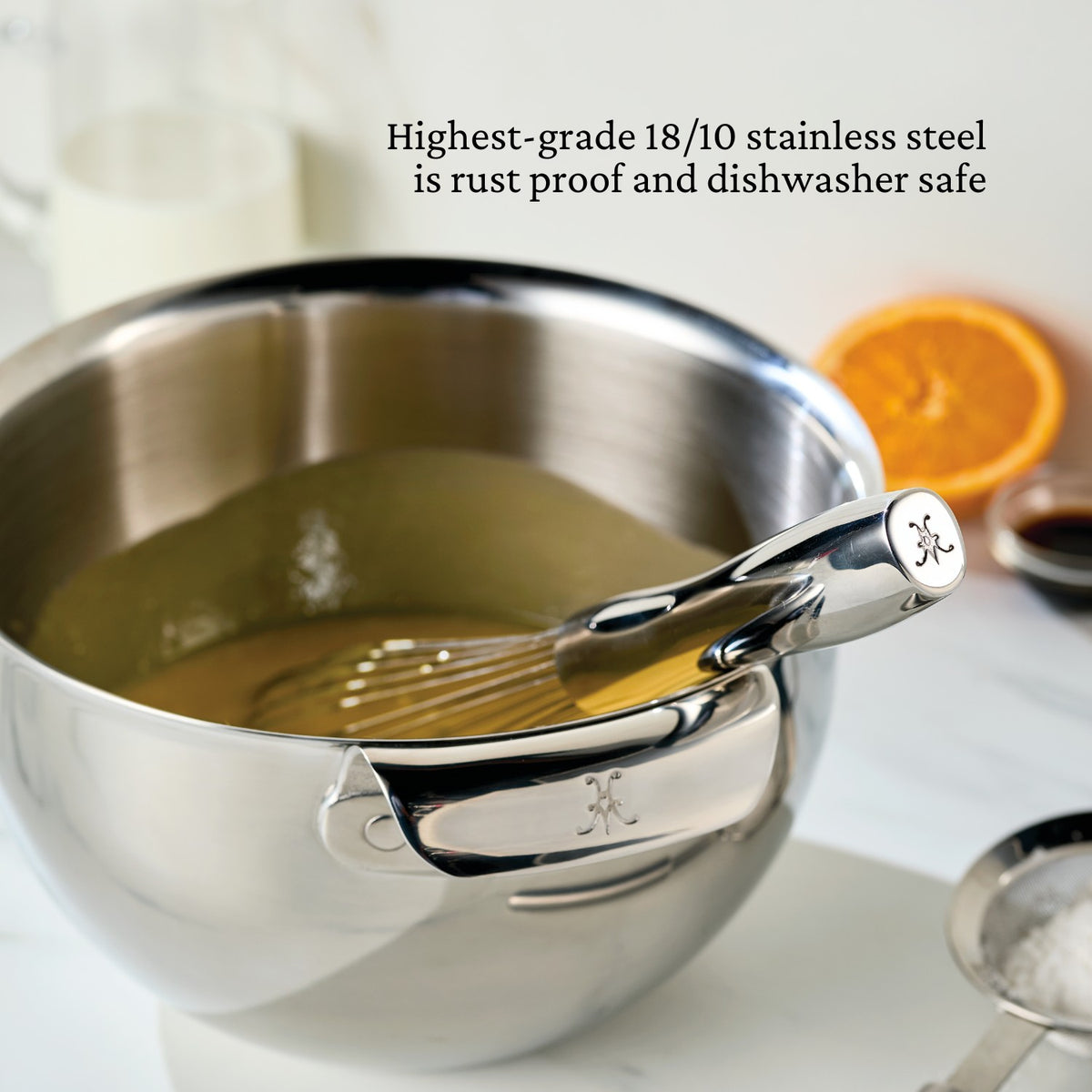 A Hestan Provisions mixing bowl holds the batter, with a Balloon Whisk inside. Made from high-grade 18/10 stainless steel, its both rust-proof and dishwasher safe. An orange slice and cup are gently blurred in the background.