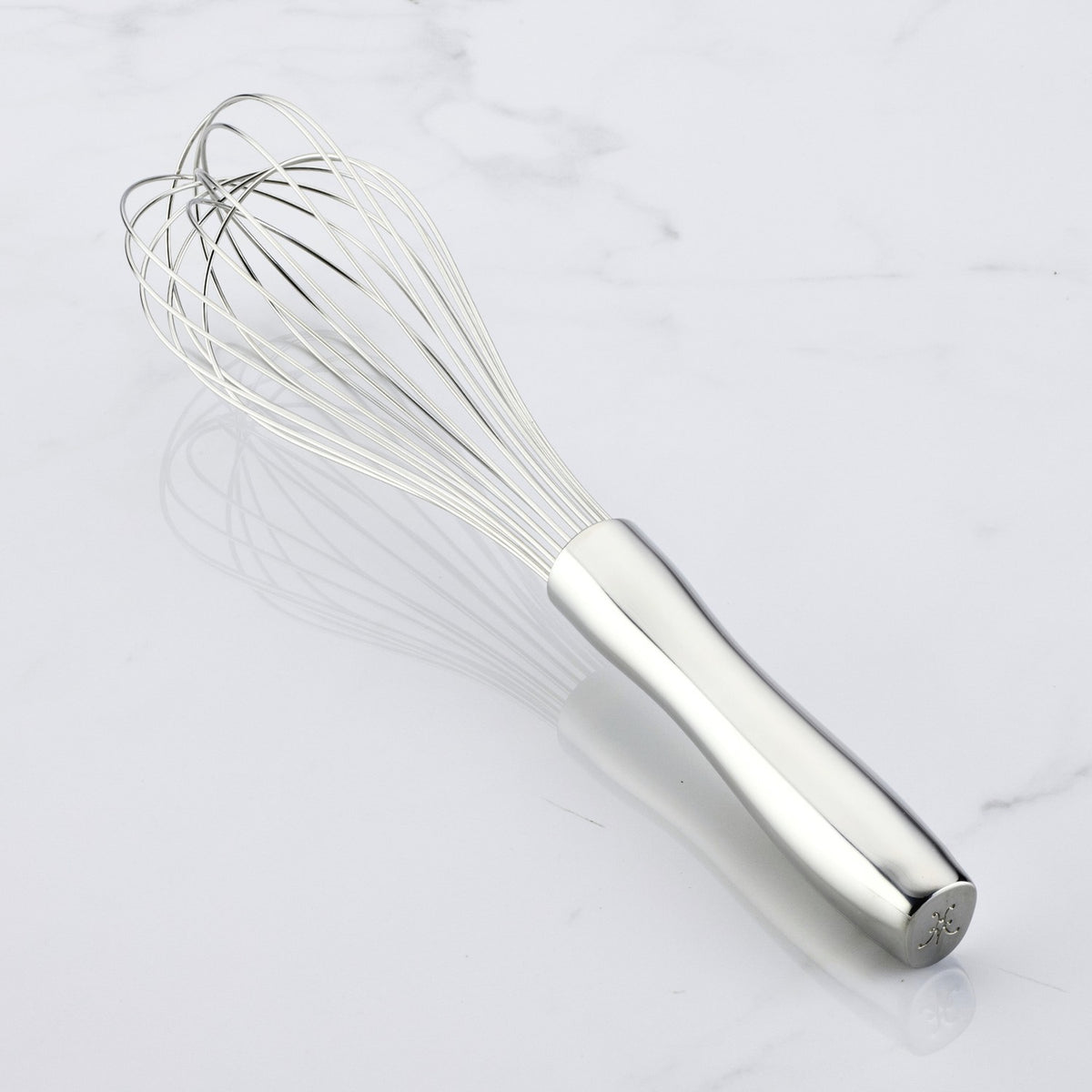 A Hestan Provisions Balloon Whisk, featuring a polished ergonomic handle and gracefully converging wires, rests on a smooth white surface with delicate shadows dancing around it.