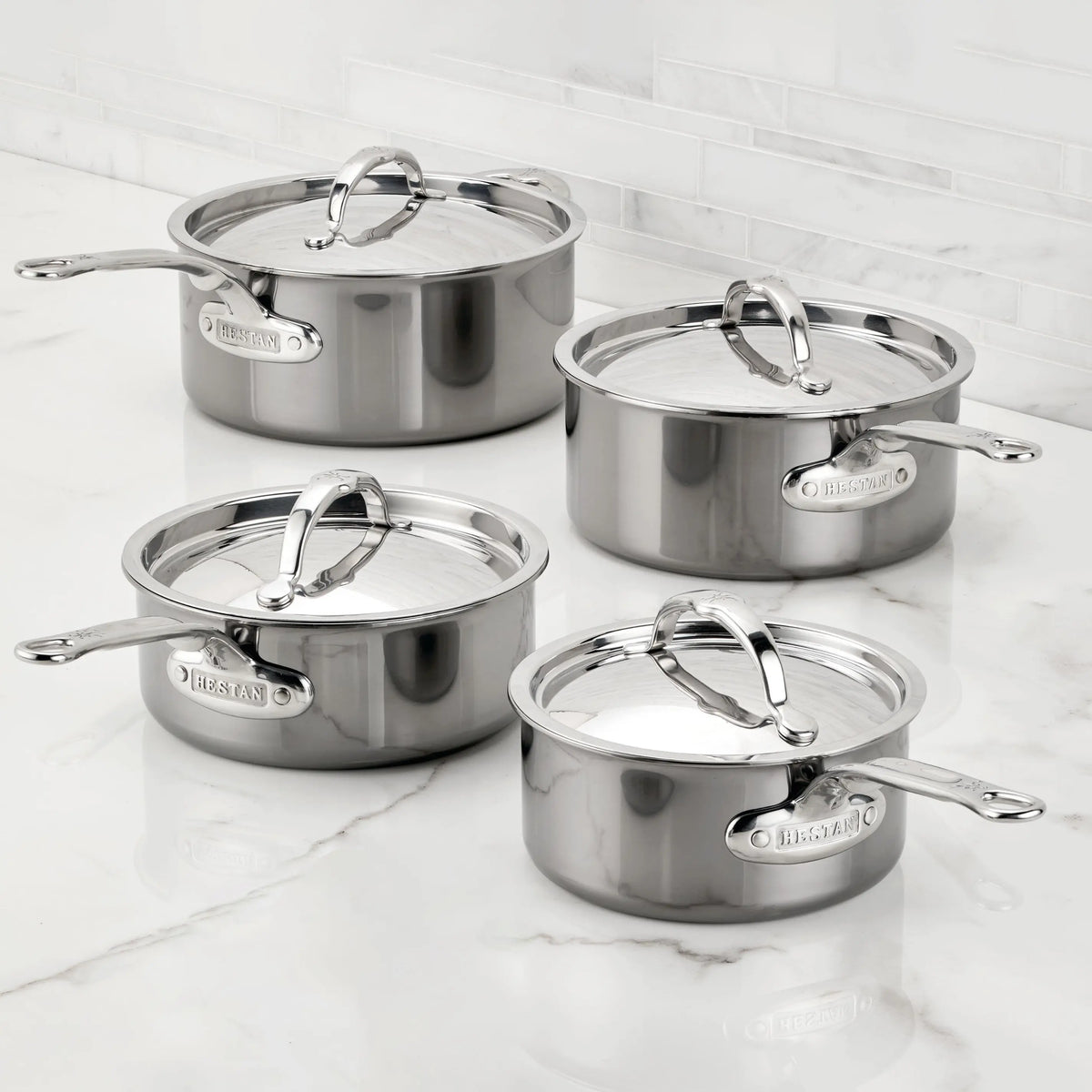 Four NanoBond Titanium Saucepans with lids are neatly arranged on a marble countertop. Each saucepan features a shiny finish, sturdy handles, and is metal utensil safe. A sleek white tiled wall provides the perfect backdrop.